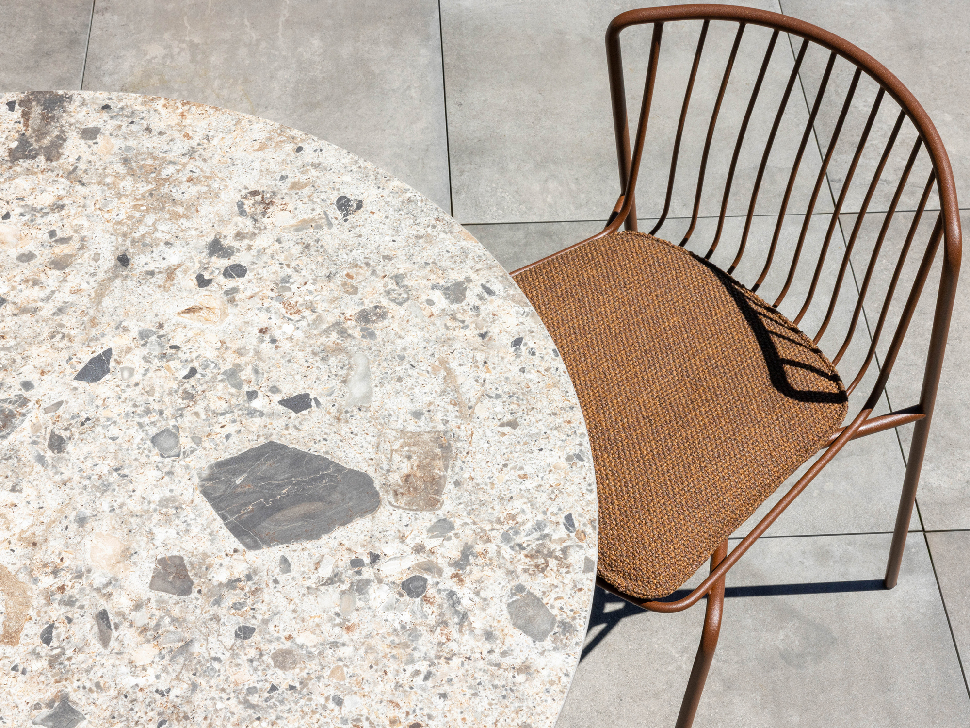 A round, speckled stone table is partially visible next to an empty brown metal chair with a woven seat, set on large square gray floor tiles. The scene is well-lit by natural sunlight.