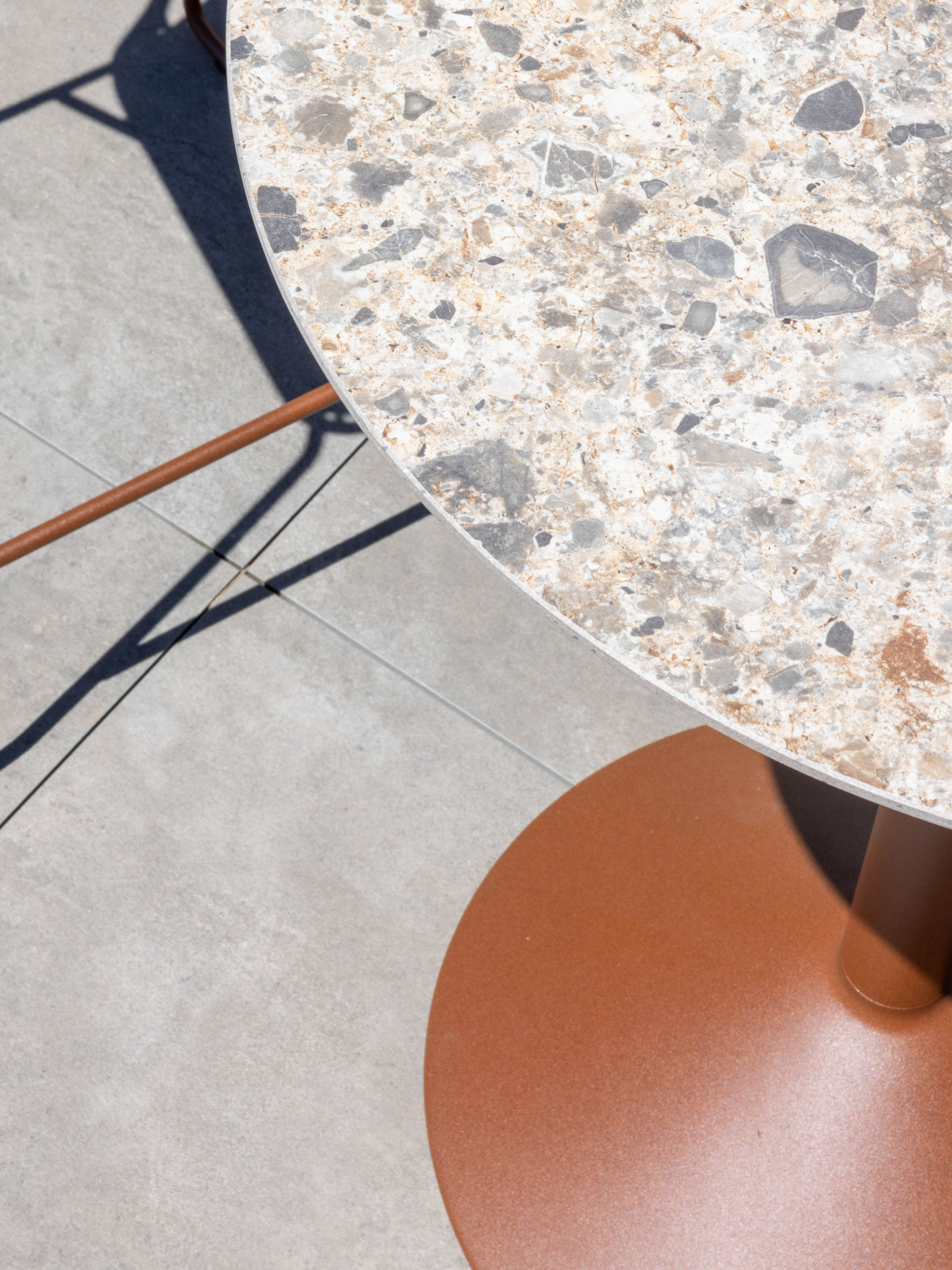 A round terrazzo table with a brown metal base stands on a light gray tiled floor. Sunlight casts sharp shadows from the table and nearby chair, emphasizing the textures and geometric shapes.