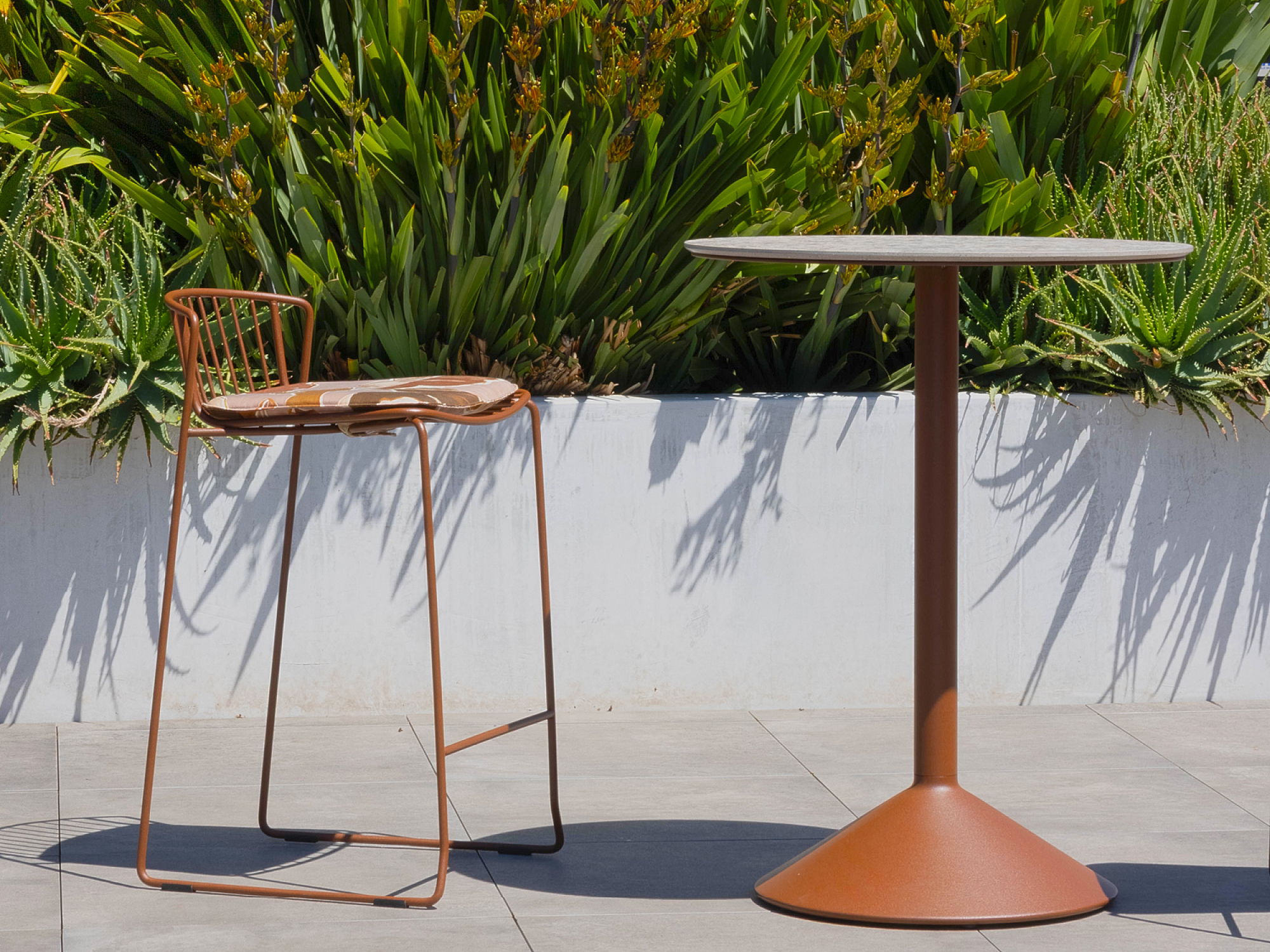 A rust-colored metal bar stool with a cushion and a matching round table stand on a tiled patio, set against a white planter filled with lush green plants.