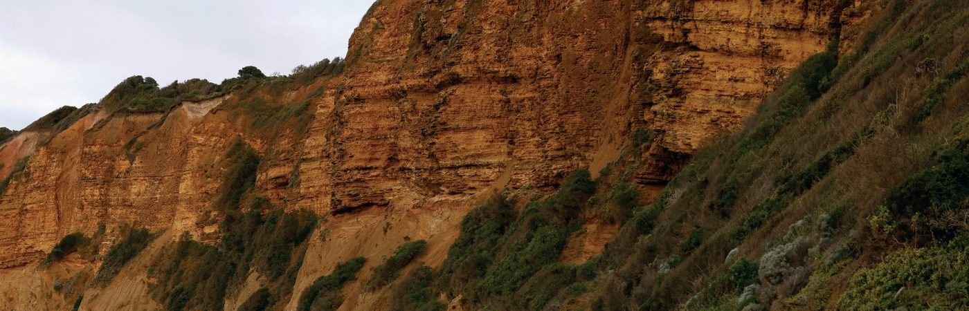 Steep, reddish-brown cliffs with patches of green vegetation rise above a sandy beach under a cloudy sky.