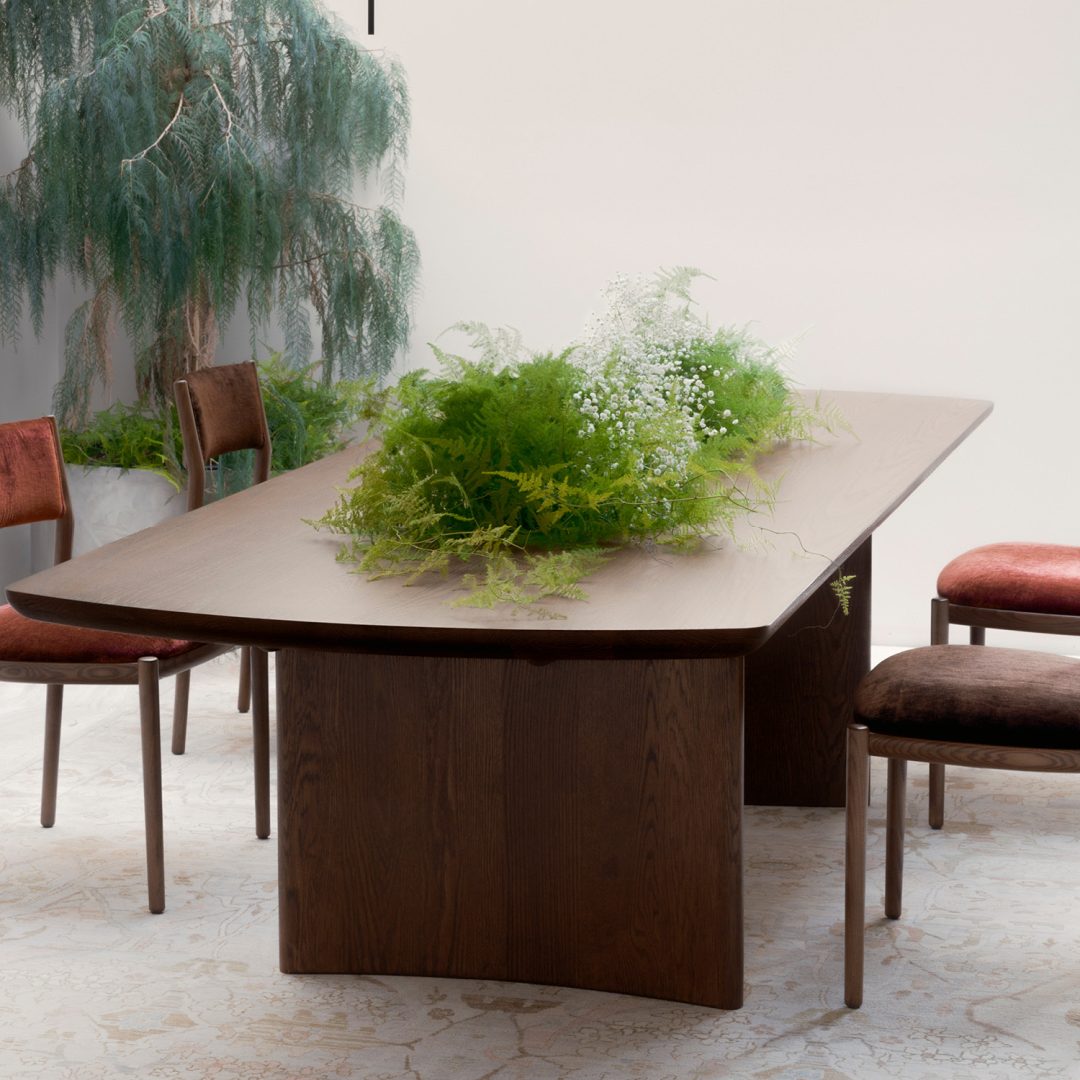 A modern dining table with a dark wood finish is surrounded by four velvet chairs in shades of red and brown. Lush green plants and white flowers form a centerpiece on the table; a tall potted plant is in the background.