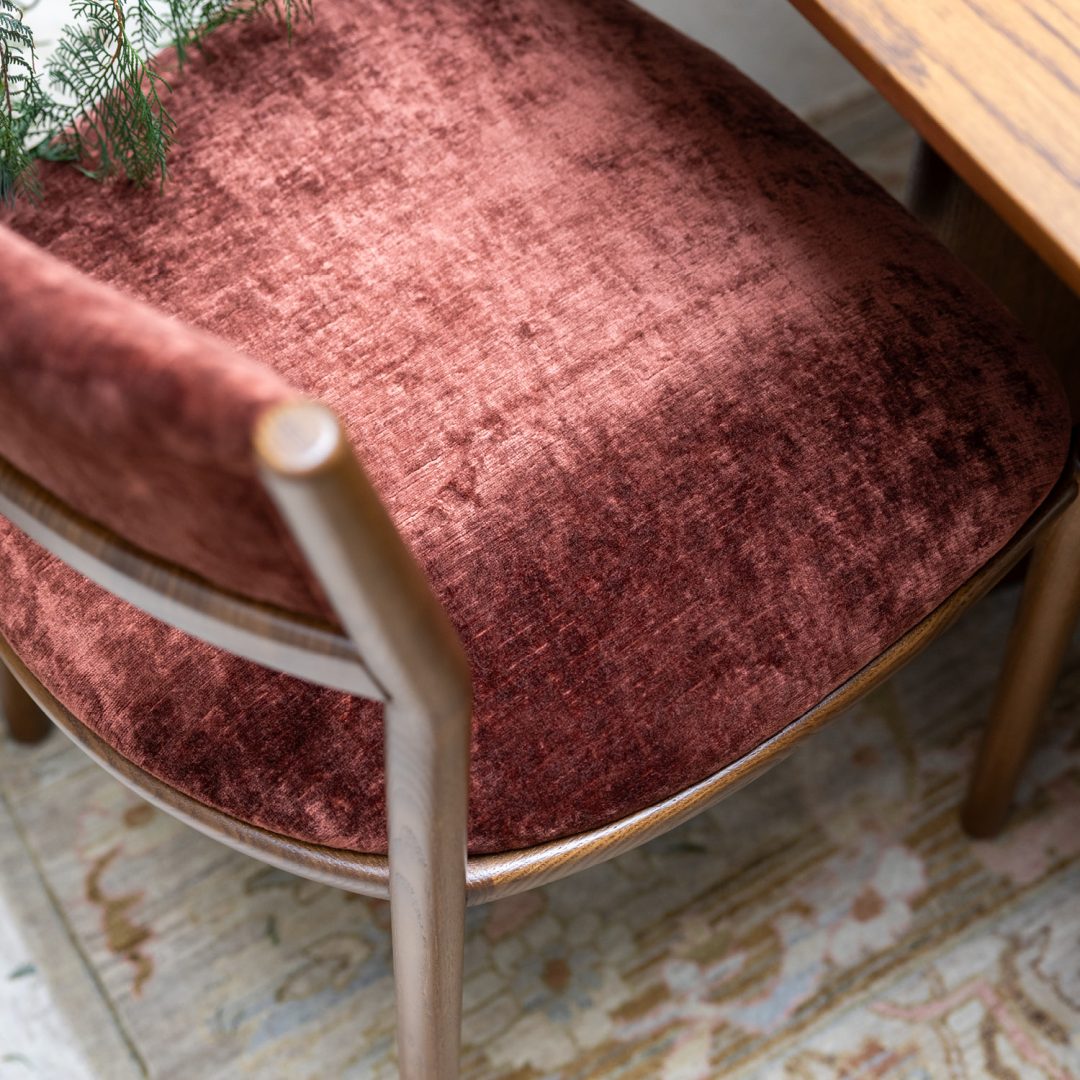 A close-up, top-down view of a wooden chair with a plush burgundy velvet seat and backrest next to a wooden table, partially over a patterned rug. A green leafy plant hangs into the frame from the upper left corner.