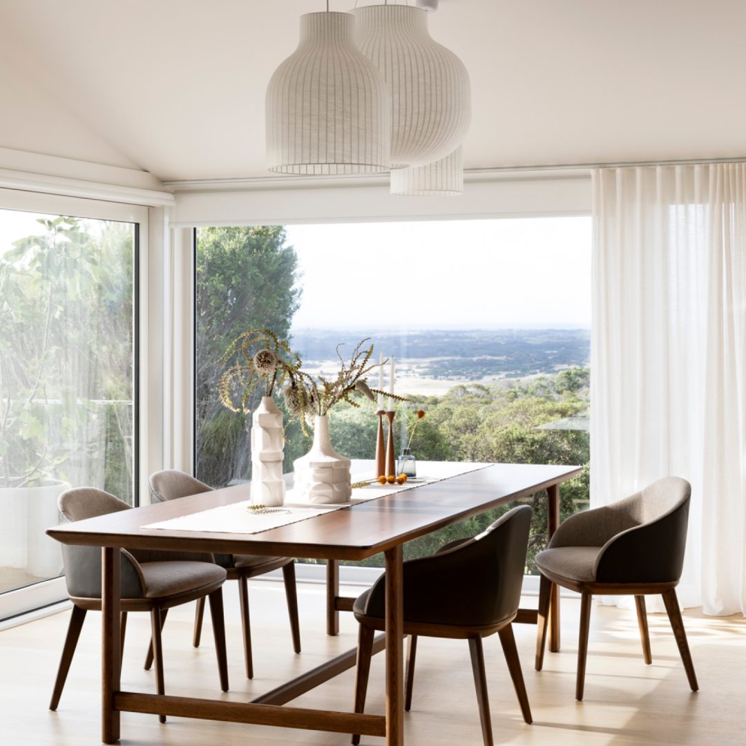 Bright, modern dining room with a wooden dining table, four brown chairs, minimalist decor, large windows, white pendant lamps, and a scenic outdoor view. Light wood floor and sheer curtains enhance the airy feel.
