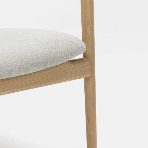 A close-up view of a modern wooden chair with a minimalist design. The chair features a light beige cushioned seat and slender wooden legs and armrests. The background is plain white, emphasizing the chair's clean lines and natural wood finish.